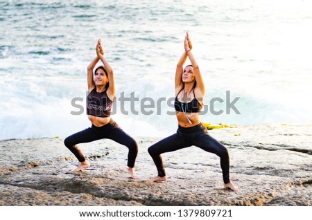 Woman yogi in a dark suit practicing yoga concept, standing in the exercise of Sumo Squat, Goddess pose, working, beach background with waves