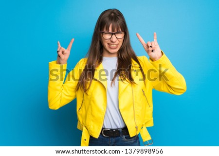 Young woman with yellow jacket on blue background making rock gesture