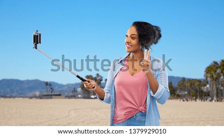 technology, gesture and people concept - happy african american woman taking picture by smartphone on selfie stick and showing peace hand sign over venice beach background in california