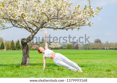 Beautiful woman is practicing yoga, doing Camatkarasana pose near blossom tree at the park.