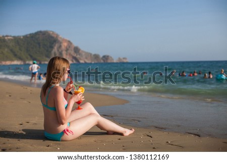 Beautiful woman with fruit cocktail on a tropical beach. Long legged girl takes sunbathe on sunbed, near the sea. 