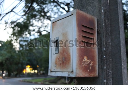 The old metallic box is hanging on cement pole in public park. Outside of box has brown rust.