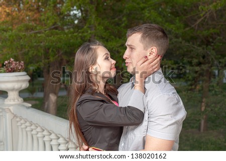 A lovely young couple hugging in the park, against the background of a romantic fence with a flower vase of French style. She holds his face with her palms and gently looks at him.