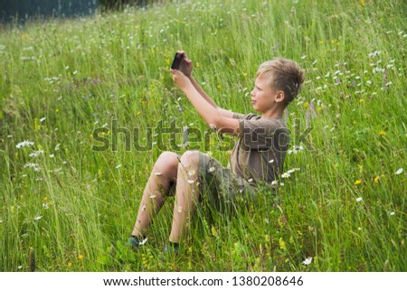 Cute young caucasian kid taking selfie of himself sitting in beautiful green meadow at countryside landscape on summer warm day. Boy using digital camera of smartphone for photography.