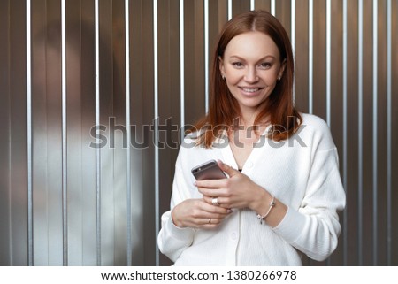 Positive young woman with appealing look, has satisfied facial expression, dressed in white clothes, reads article on webiste, looks gladfully, receive feedack from business colleague, uses wifi