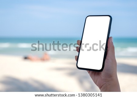 Mockup image of woman's hand holding black mobile phone with blank desktop screen by the beach and sea 