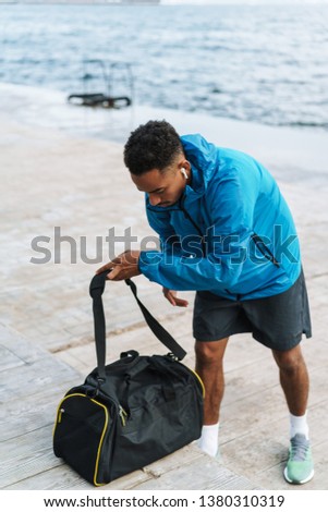 Picture of a handsome young african sports man outdoors at the beach sea walking with bag listening music with earphones.