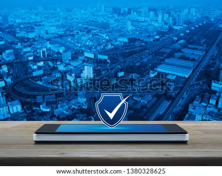 Security shield with check mark icon on modern smart mobile phone screen on wooden table over city tower, street, expressway and skyscraper, Technology internet cyber security and anti virus concept