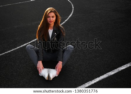 Glamorous modern young female athlete in black stylish jeans in a black leather jacket in fashionable leather white sneakers on an open basketball court on a spring Sunny day. Nice beautiful girl.
