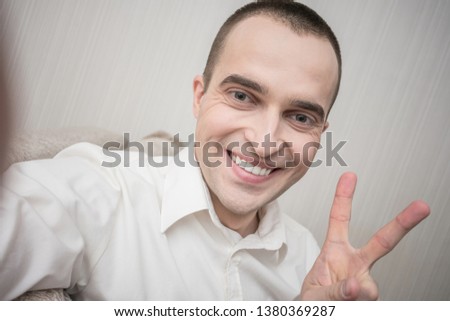 Attractive young man in white shirt smiling for selfie, shows sign peace, sitting at home on the couch, front view