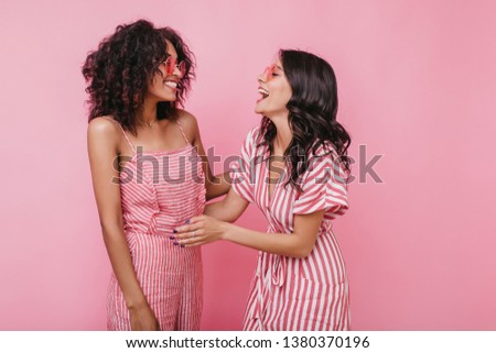 Girl with amazing smile tells her friend funny story. Indoors portrait of models with tanned skin on pink background