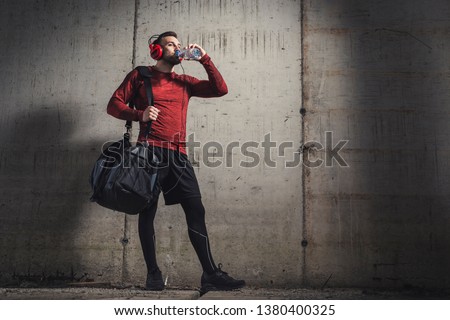 Muscular athetic man wearing sportswear and carrying a gym bag, listening to the music and drinking water after a hard workout Royalty-Free Stock Photo #1380400325
