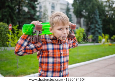 Closeup portrait of cute white kid standing in park alone and holding bright green plastic mini cruiser in hands. Horizontal color photography.