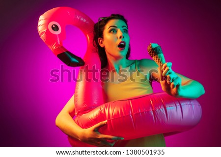 Happy young woman in a rubber flamingo and sunglasses with an icecream on trendy pink neon studio background. Female portrait. Concept of human emotions, facial expression, summer holidays or weekend.