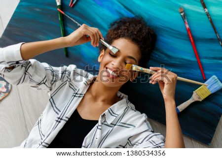 Top view of happy  painter   woman lying on canvas  with  brushes  in hands.  Dreaming  and relaxing after productive work. 
