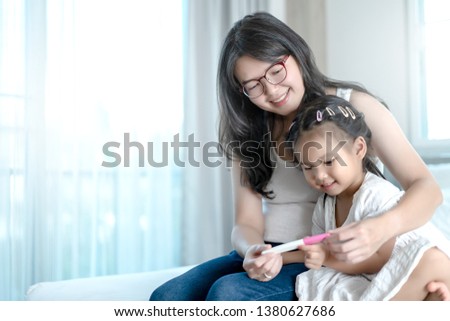 Asian attractive mother smiling and shows the result positive on pregnancy test tools to her first daughter on the bed in their bedroom. Her daughter feels excited and glad.