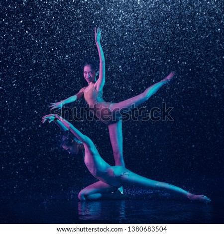 Look. Two young female ballet dancers under water drops and spray. Caucasian and asian models dancing together in neon lights. Ballet and contemporary choreography concept. Creative art photo.