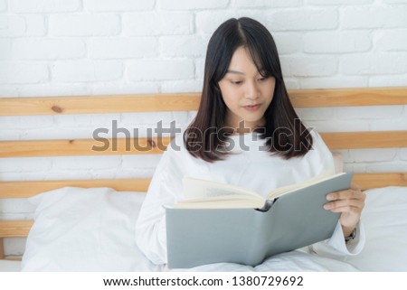 Asian woman reading a book and smiling as she sits in bed.