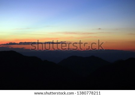 Dark blazing misty sunset landscape of dark blue-orange cloudy sky with silhouette of mountains, shown as a background. Beautiful summer sunset viewing from the peak of forest mount.