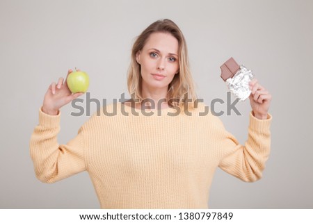 Beautiful young blond girl holds a bar of chocolate and a green apple around her face, she can not make a choice. She is dressed in a yellow sweater. poses against a white background.facial expression