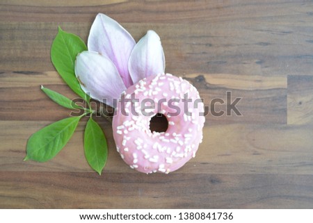 A pink donut with white sprinkles and magnolia petals lie on a dark wooden surface