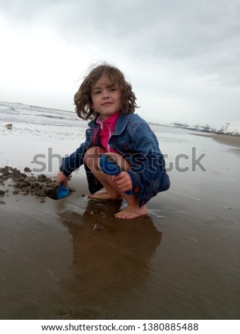 little girl in a denim jacket playing with wet sand near the sea, in cold weather, on the beach against the background of the port of Valencia. Picture taken April 23, 2019, Malvarrosa beach, Valencia