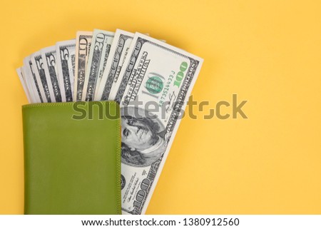 Several hundred dollar bills protrude from a stylish green leather wallet on a bright yellow background. Top view, close-up.