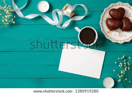 Wedding invitation concept. Blank paper card with daisy flowers, heart-shaped chocolate and coffee cup on green wooden background. Tender women's desk, flat lay style composition, top view, overhead.
