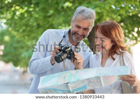 Two mid adult tourists visiting a foreign city. Walking through the park, woman is holding a map and man is showing pictures on a digital camera