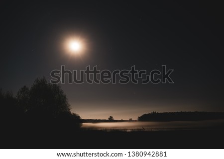 A full moon illuminating the valley flooded in the mist. In the distance you can see the contour of the forest. Beautiful clean, starry sky. Long exposure at night