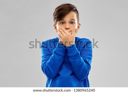 childhood, expressions and people concept - shocked little boy in blue hoodie closing his mouth by hands over grey background