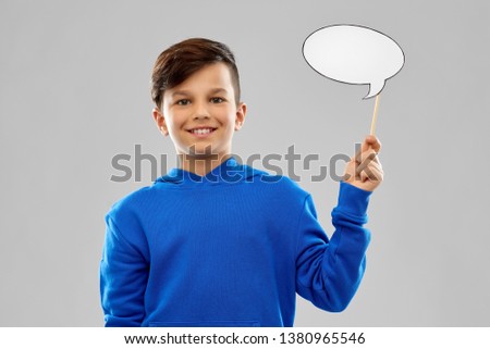 party props, photo booth and communication concept - smiling boy in blue hoodie holding blank speech bubble over grey background Royalty-Free Stock Photo #1380965546