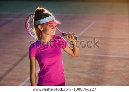 Handsome young girl in stylish cap and sportswear with racket on shoulder on the sunshine background. Female tennis player on outdoor court at the sunset.