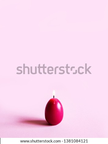 Traditional Easter decor. Bright burning paraffin candle in the shape of colorful egg on soft pink background.