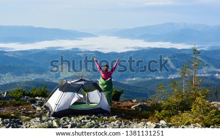 Camping on the top of mountain in the morning. Back view of young hiker woman in pink sweater and sleeping bag standing beside tourist tent, holding hands lifting up. On foggy mountains background.