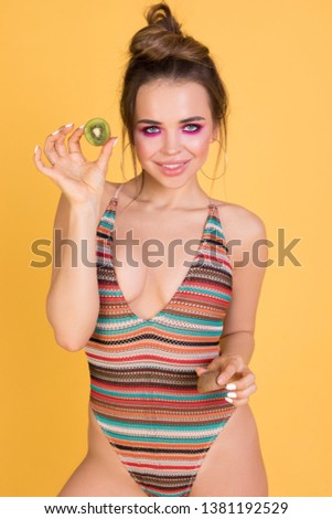 Emotional girl in a swimsuit holding a kiwi on a yellow background.