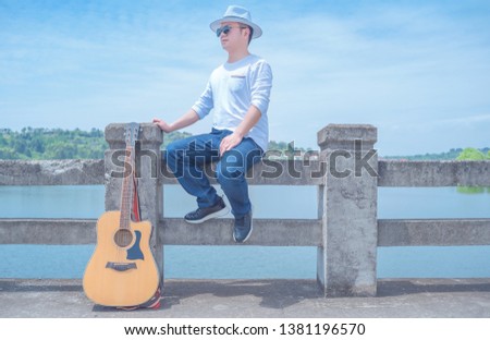 In a hot summer , a young literary youth who wearing a white T-shirt and jeans and a white Panama hat, plays the guitar alone on the dam under the blue sky.