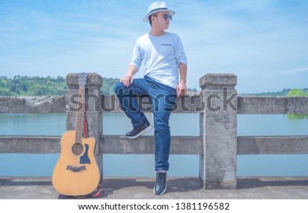 In a hot summer , a young literary youth who wearing a white T-shirt and jeans and a white Panama hat, plays the guitar alone on the dam under the blue sky.