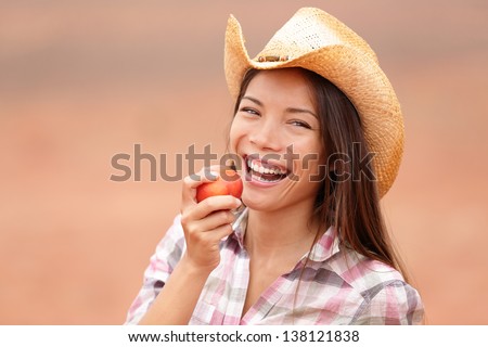 American cowgirl eating peach or nectarine fruit smiling and laughing happy wearing cowboy hat outside. Healthy eating concept with beautiful young mixed race Caucasian Asian female model outdoor.