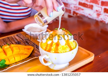 Woman hand pouring sweetened condensed milk on ice Bingsu (Korean dessert style) refreshing dessert with mango fruit and sticky rice.