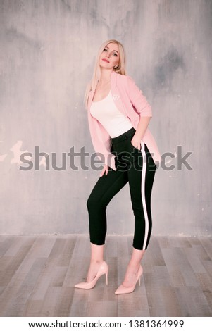 Attractive positive middle-aged blond woman wearing pink jacket and pants with trouser stripes with a beautiful smile posing against a receding wall looking directly at the camera