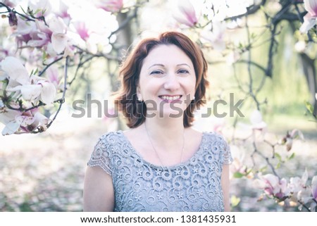Happy middle aged Armenian woman in an elegant dress under the blooming magnolia tree. Smiling, looking at the camera. Toned, selective focus.