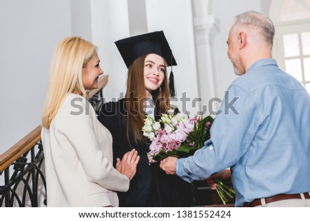 happy father giving flowers to cheerful daughter in graduation cap  Royalty-Free Stock Photo #1381552424