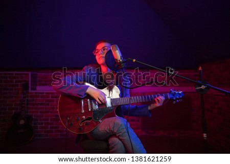 A young woman in glasses playing guitar and singing in neon lighting