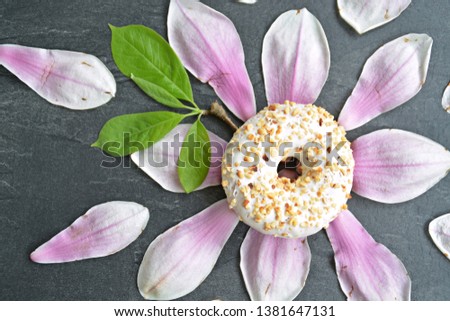 A white glazed donut with brittle splinters lies on a dark surface with petals of a magnolia