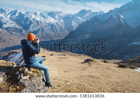 Portrait of a bearded male photographer in sunglasses and a warm jacket with a backpack sits on a big stone and takes pictures on camera against the background of snow-capped mountains