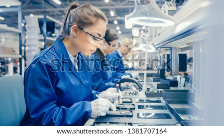 Woman Electronics Factory Worker in Blue Work Coat and Protective Glasses is Assembling Smartphones with Screwdriver. High Tech Factory Facility with more Employees in the Background. Royalty-Free Stock Photo #1381707164