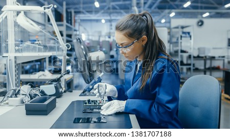 Young Female Blue and White Work Coat is Using Plier to Assemble Printed Circuit Board for Smartphone. Electronics Factory Workers in a High Tech Factory Facility. Royalty-Free Stock Photo #1381707188