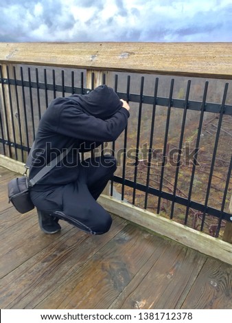 Photographer man bending down to shoot a picture wearing a black hoodie and jeans on a wooden bridge overlooking woods