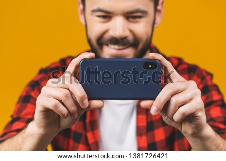 Excited bearded man in plaid shirt playing on smartphone isolated over yellow background.